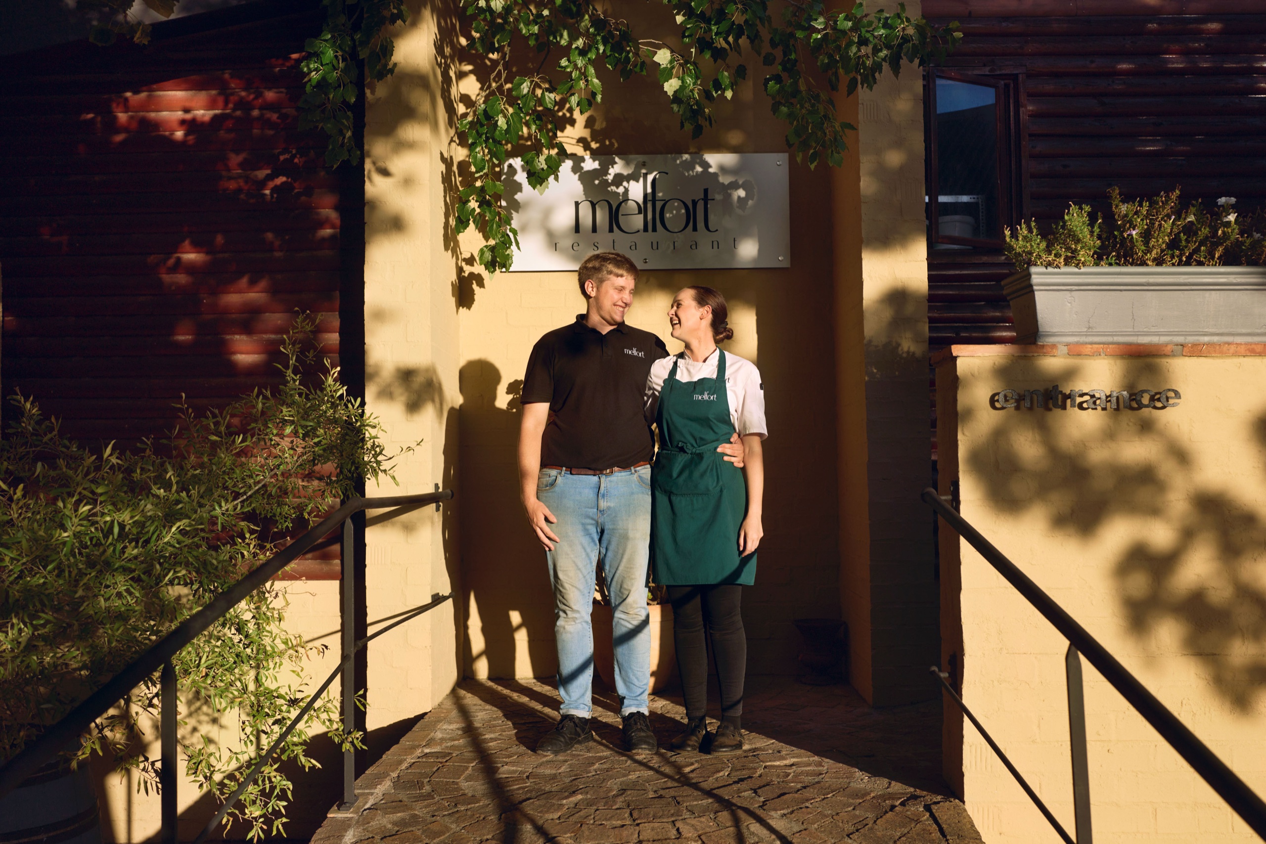 Two restaurant owners stand arm in arm at the entrance of Melfort in warm evening light