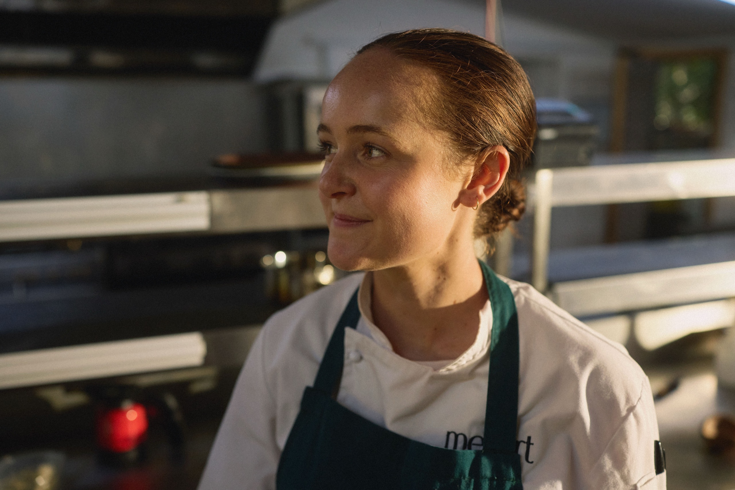 A chef in a green apron stands in the restaurant kitchen, lit by warm evening light
