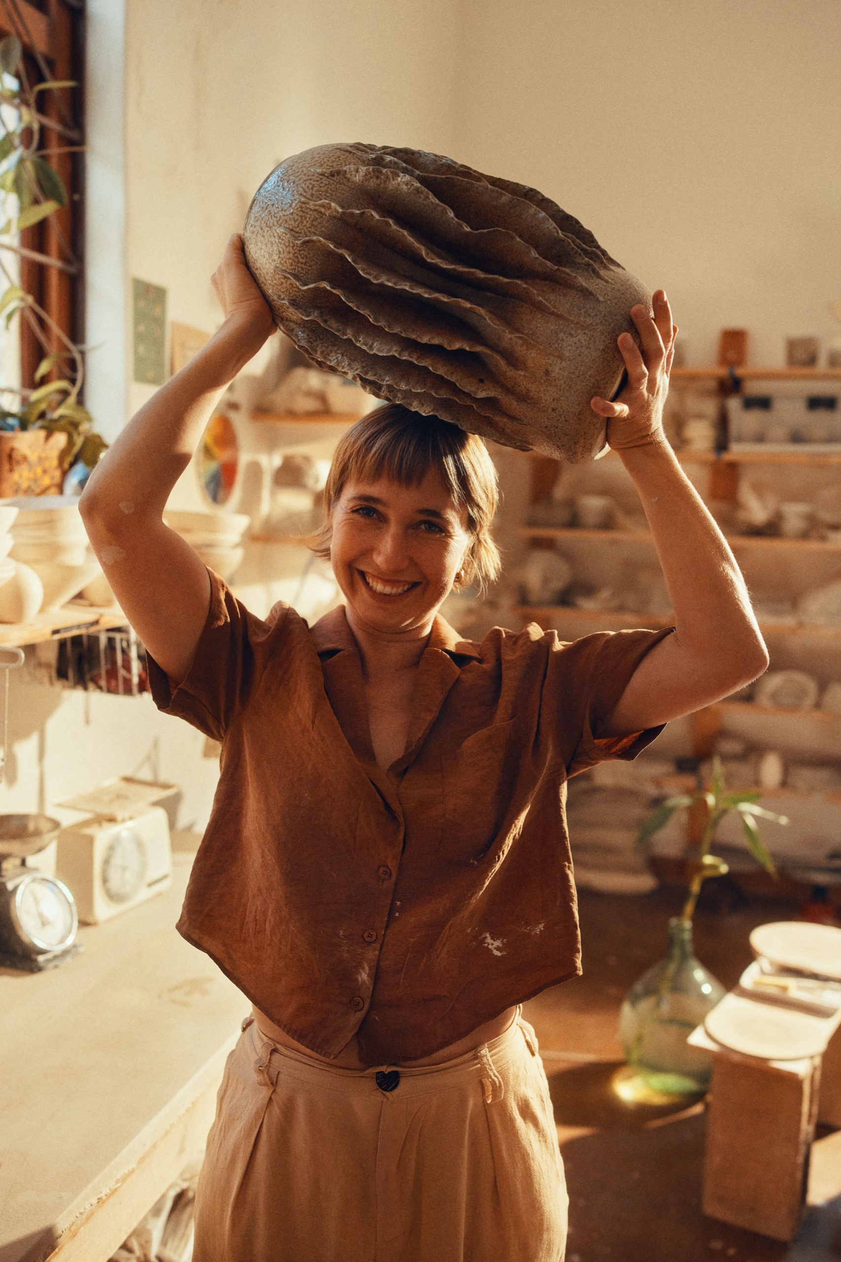 A maker holds a large sculptural ceramic vessel above her head in a sunlit studio