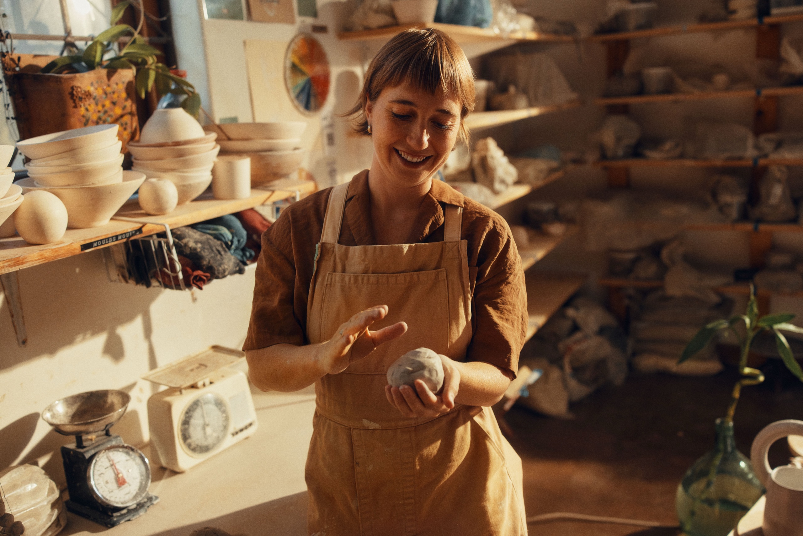 A ceramicist holds a small clay form in a bright, daylit studio with scales and shelves behind her