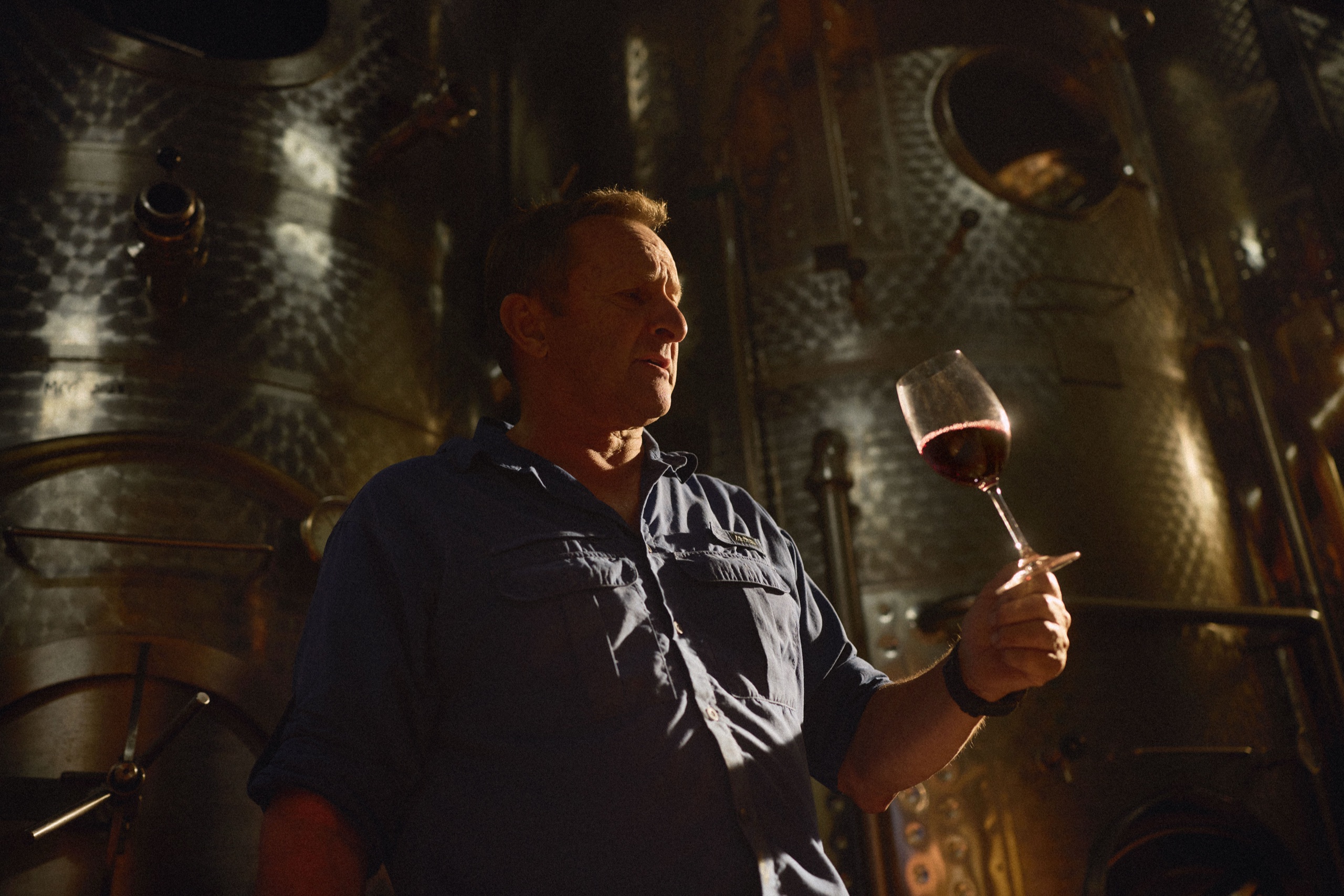 A winemaker holds a glass of red wine up to the light beside a copper still